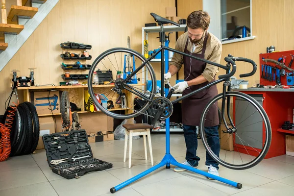 年轻英俊的技工穿围裙修理自行车 当听音乐在耳机 内部现代车间在背景 — 图库照片
