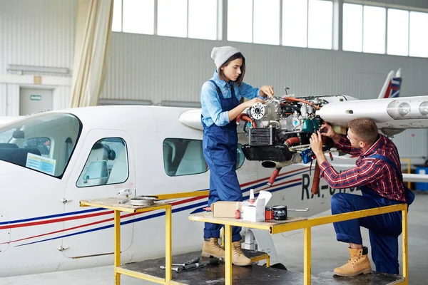 Airplane service crew repairing plane in hangar:  two young mechanics, man and woman, fixing jet plane turbine