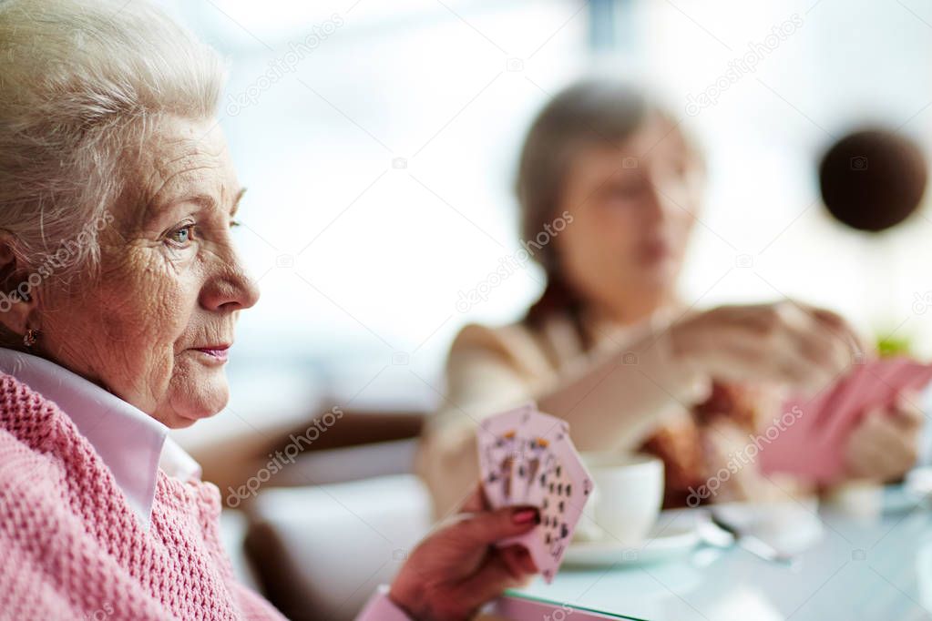 Profile view of pensive elderly woman with deep blue eyes sitting at table and playing cards with her female friend