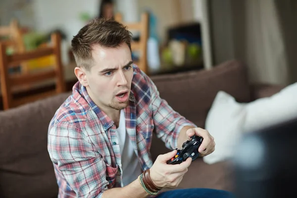 Portrait of emotional adult man playing video game holding wireless controller and  opening mouth while sitting on couch at home