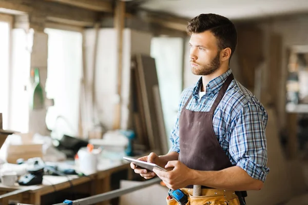 Dalgın genç woodworker dijital tablet atölye, bel, dikey pencereden dışarı bakarken elinde tutan