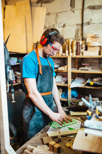 Портрет сфокусированного молодого человека, работающего в столярной мастерской, резки деревянных деталей в защитных очках и наушниках

