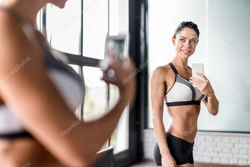 Portrait of proud muscular  woman boasting her fit figure and slim waist taking sefie in gym  mirror and posing after working out