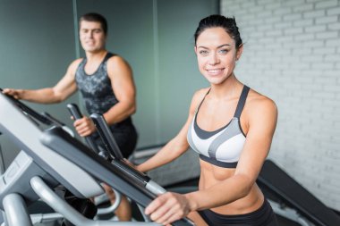 Eliptik sonraki her iki gülümseyen kameraya modern spor olarak çalışma sırasında adam arayan, sığacak şekilde makinasıyla egzersiz güzel sportif esmer kadın portresi