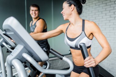 Uygun adamın yanında eliptik makinesi kullanma ve ona modern spor olarak çalışma sırasında gülümseyen egzersiz güzel sportif esmer kadın portresi