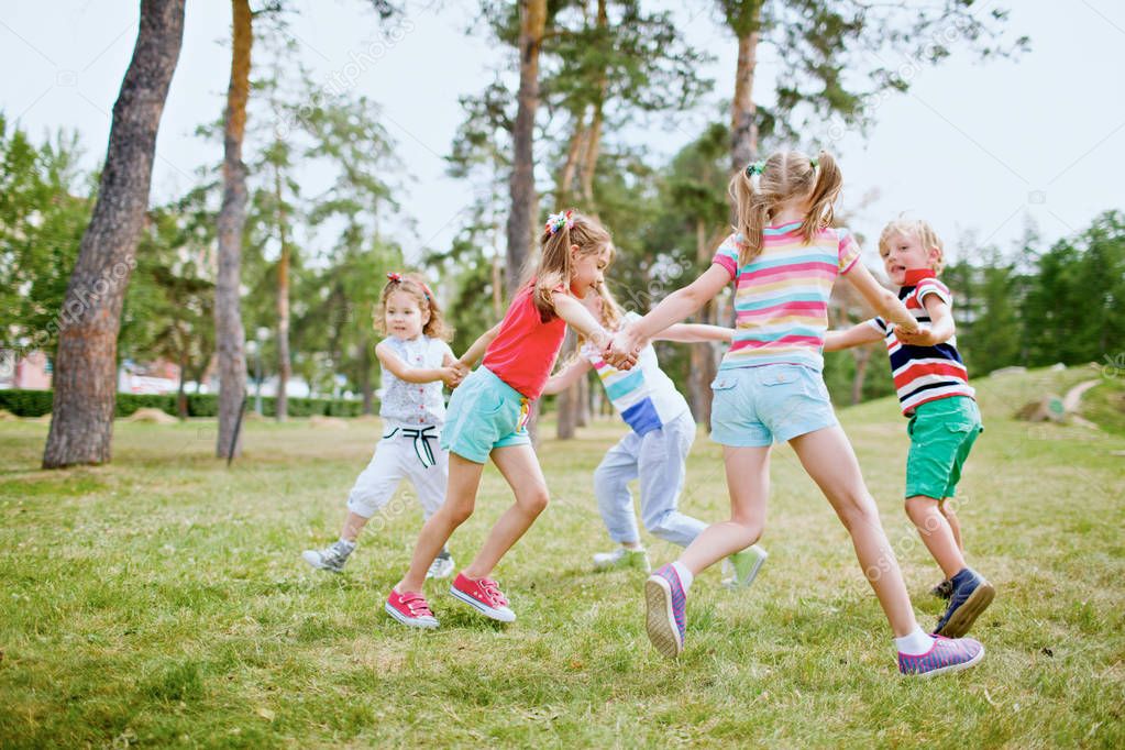 Group of children holding hands and dancing in circle on green lawn in park on beautiful summer day