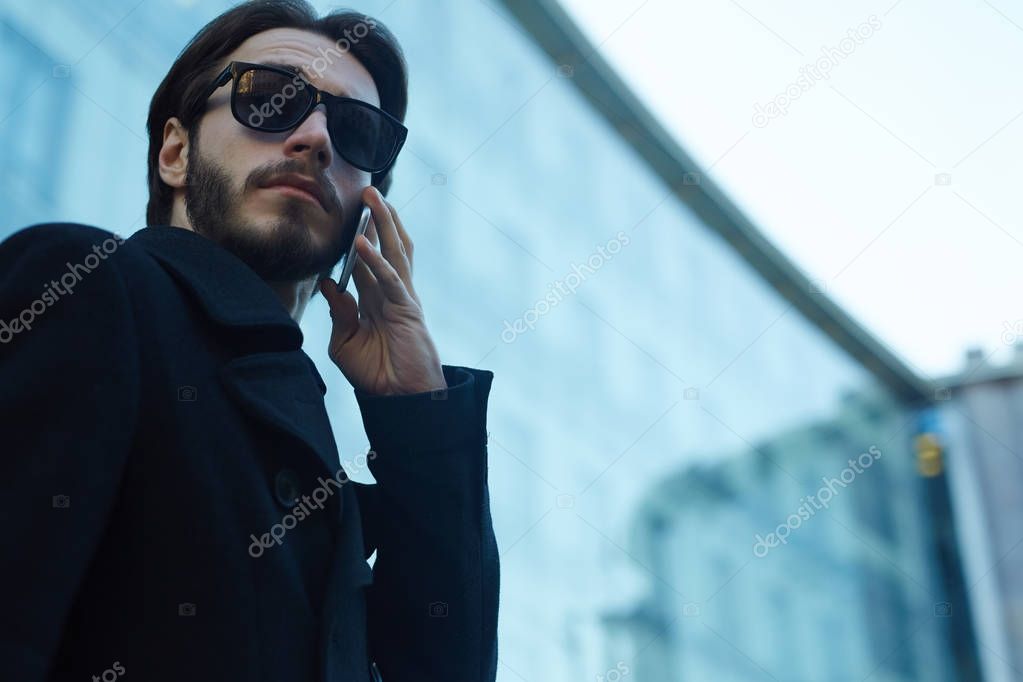 Low angle portrait of handsome modern man wearing sunglasses and black coat making phone call outdoors in streets of business city