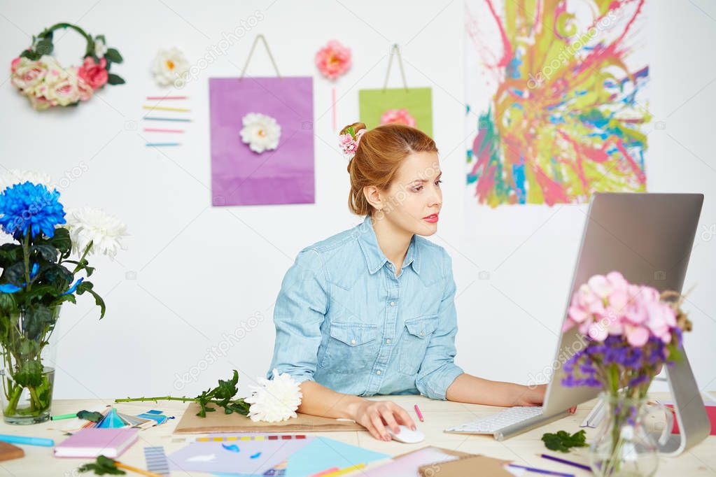 Concentrated graphic designer creating logo for client while sitting at messy office desk, white wall decorated with flowers and abstract painting on background