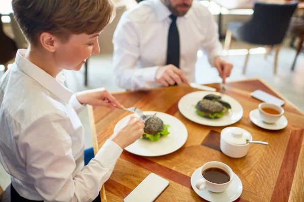 在现代咖啡馆吃午餐的时候 她的男同事坐在她旁边 面带微笑的漂亮的女商人享受美味的汉堡 — 图库照片
