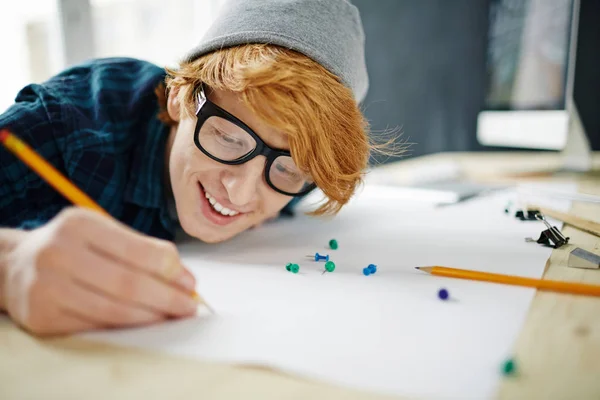ビーニー帽子と鉛筆デスクに近い傾いたと元気よく楽しんで仕事を笑顔でメガネを身に着けている若い創造的な赤髪の男の肖像 — ストック写真