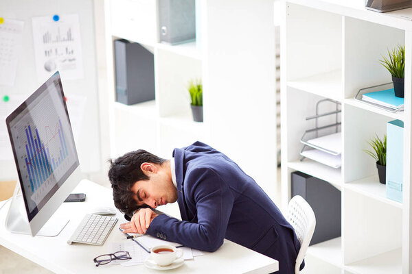 Перегруженный работой и уставший молодой менеджер в стильном костюме спит на столе перед компьютером, интерьер современного офиса на заднем плане
