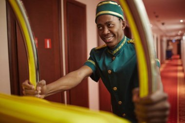Otel odalarında koridorda çanta sunma Bagaj kart bastırmak gülümseyen genç belboy Afrika etnik köken, çalışan bir portresi