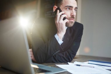 Dizüstü bilgisayar kullanarak ve mercek parlaması ile siyah arka plan karşı cep telefonla konuşurken ofiste çalışan modern sakallı işadamı portresi