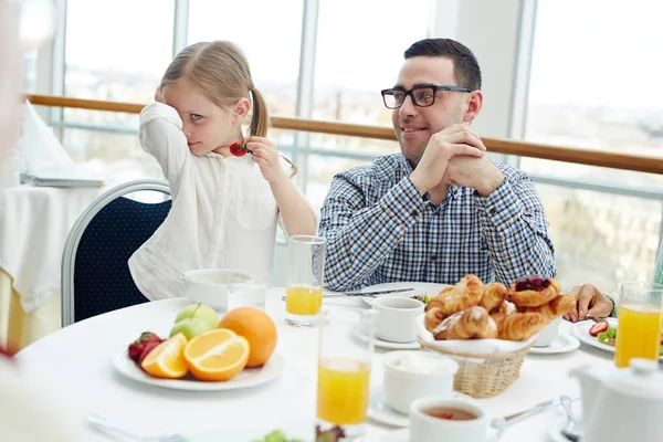 いたずらな女の子の若い男が彼女を見ている間テーブルでイチゴを食べる — ストック写真