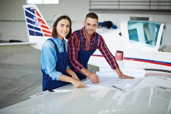 Two modern aircraft engineers, man and woman, looking at camera while working with plane blueprints in airport hangar