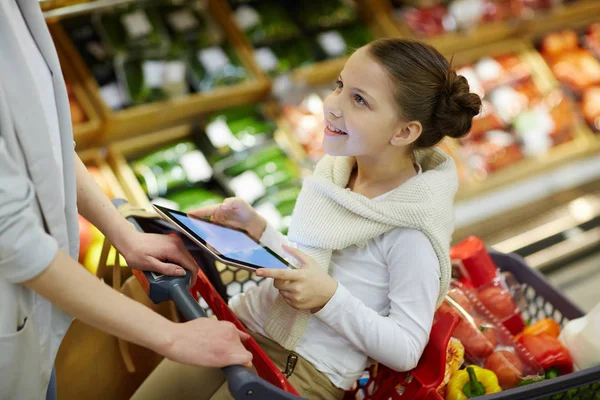 ショッピングカートと彼女自身を心に抱く彼女の両親食料品のスーパーで買い物しながらデジタル タブレットに座っている笑顔の少女の肖像画 — ストック写真