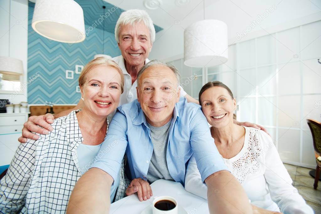Joyful elderly friends posing for photography with wide smiles while taking selfie at stylish coffeehouse, first person view