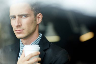 Yakışıklı genç adam kameraya kağıt bardak kahve elinde tutarken ciddi göz Görünümü penceresinden bakarak baş ve omuzlar portresi