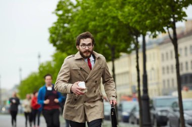 Busy man in elegant trench-coat carrying drink and briefcase while running for work after coffee-break clipart