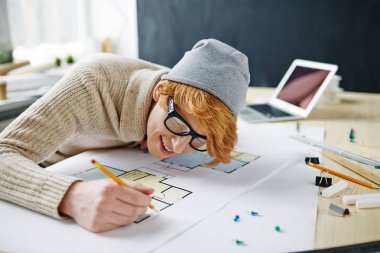 Gözlük ve çalışma rchitects office bere şapka giyen modern kırmızı saçlı adam portresi: kat planı çizim ve neşeyle gülümseyen masanın üzerine eğilerek