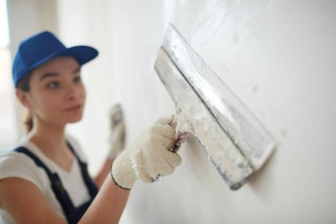 Portrait of young pretty woman on construction site: female worker plastering walls with spatula  while remodeling office clipart