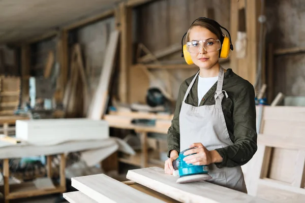 Confident fair-haired craftswoman in eye and ear protectors looking at camera while holding electric sander in hands, waist-up portrait