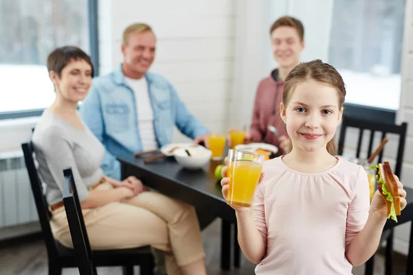 Waist 的肖像漂亮的小女孩看着相机与微笑 同时拿着橙汁的一只手和健康的三明治在其他 她的家人坐在餐桌 — 图库照片