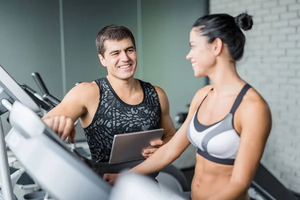 肌肉的个人教练的肖像看微笑适合的妇女使用椭圆机运动在现代健身房 — 图库照片