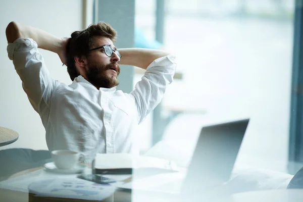 Relaxing businessman sitting in front of laptop and looking through office window