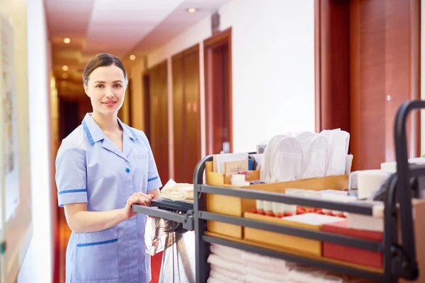 Housemaid pushing cart with clean bedclothes while walking along corridor of hotel