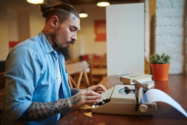 Side view portrait of stylish tattooed man using old-school vintage typewriter on wooden table in cafe, typing story