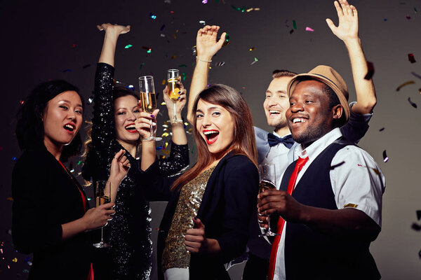Multi-ethnic group of friends clinking champagne flutes together and wishing happy New Year while having fun in night club, colorful confetti falling