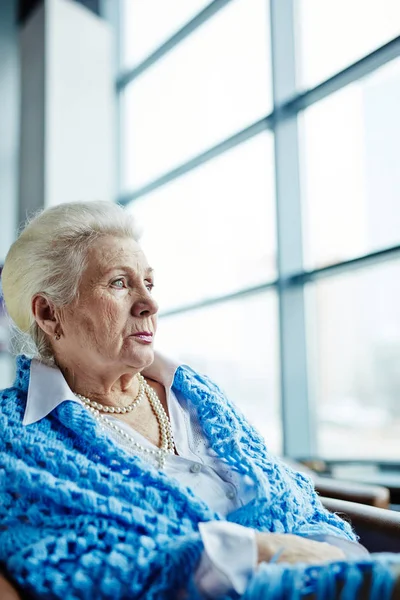Waist 美丽的老妇人与珍珠项链和矢车菊蓝色针织披肩坐在扶手椅上 看着全景窗口的肖像 — 图库照片
