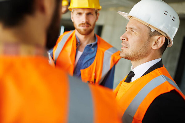Confident builder in helmet and uniform talking to his colleagues