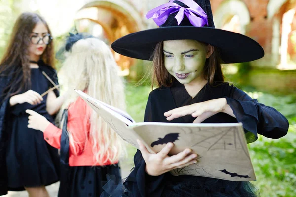 邪恶的女孩在帽子和巫婆的装束保持她的手在万圣节魔术的开放法术书 — 图库照片