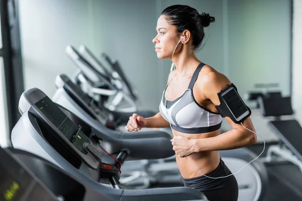体育棕褐色妇女在健身房跑步机上锻炼的肖像使用肩部智能手机支架听音乐 — 图库照片