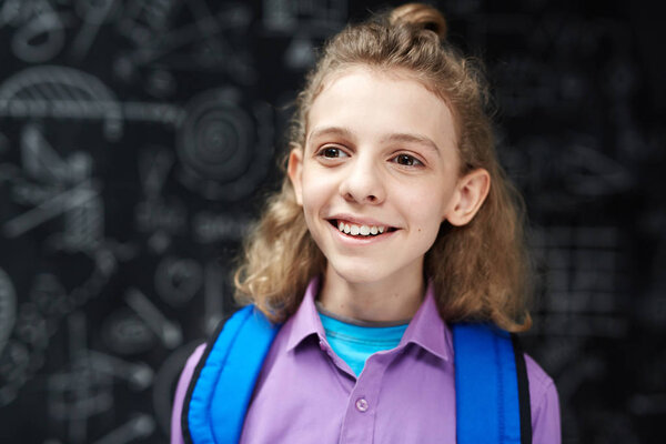 Happy boy with backpack smiling cheerfully while posing against blackboard in school