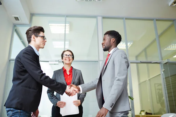 Profile view of confident business partners shaking hands as sign of successful completion of negotiations, pretty middle-aged assistant manager looking at them with toothy smile