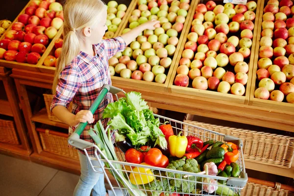 Little consumer with cart full of vegs choosing ripe apples in hypermarket