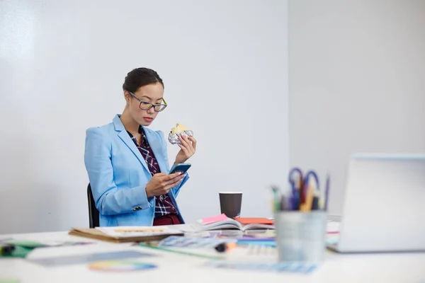 Young female with sandwich messaging in smartphone while sitting in office