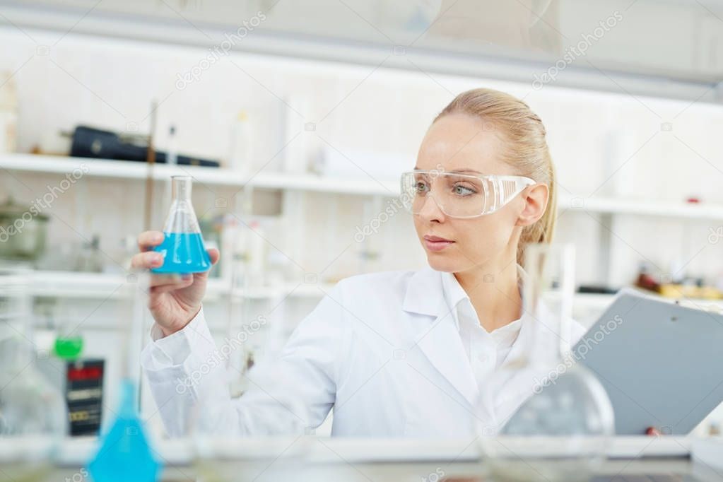 Portrait of beautiful female scientist working on research in modern laboratory holding test tubes with colored liquid and writing results