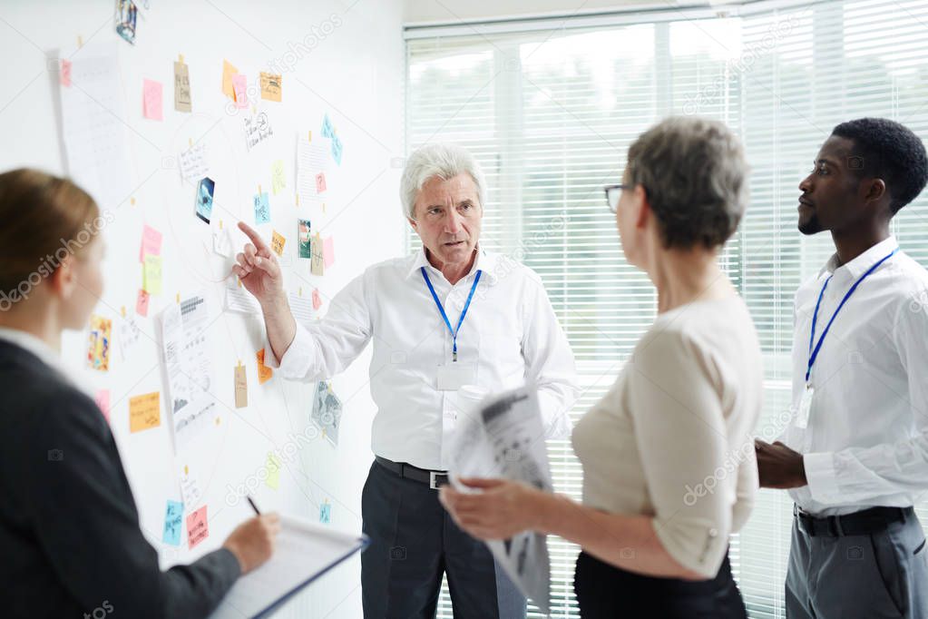 Multi-ethnic group of managers gathered together at modern boardroom and brainstorming on joint project, confident senior boss pointing at dry-erase board