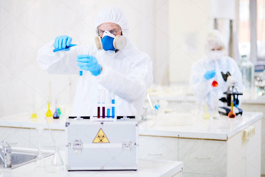 Male researcher wearing hazmat suit adding blue solution to test tube while conducting experiment at modern laboratory