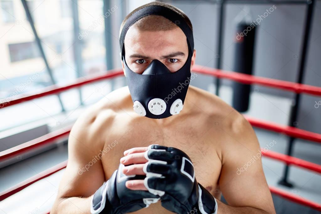 Portrait of shirtless boxer standing in boxing ring looking at camera with determination wearing training mask, ready to fight