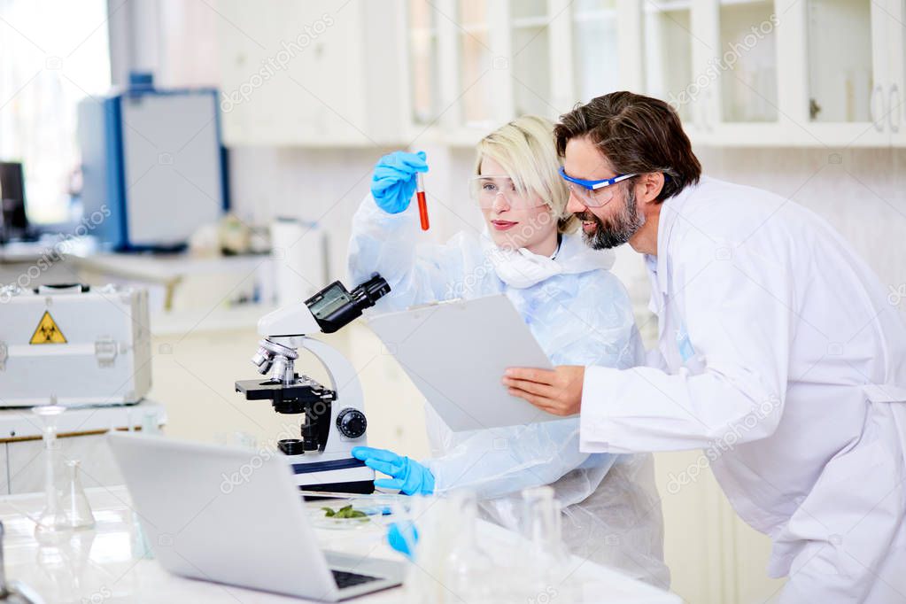 Cheerful group of microbiologists wearing safety goggles looking at test tube proudly while working together on creation of brand new cancer vaccine, interior of modern laboratory on background