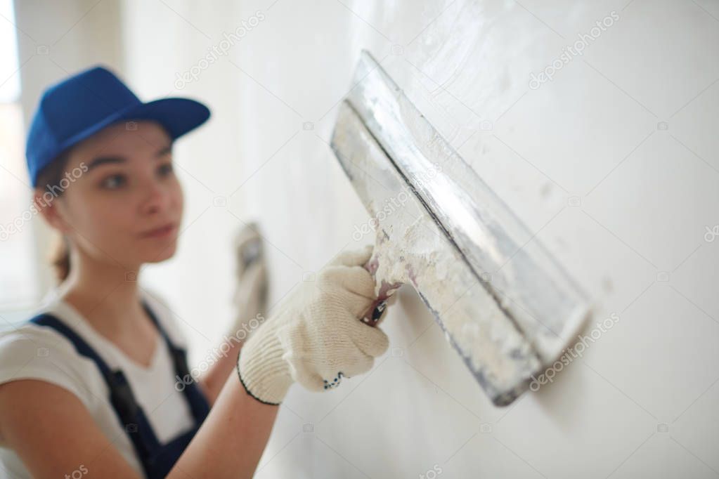 Portrait of young pretty woman on construction site: female worker plastering walls with spatula  while remodeling office