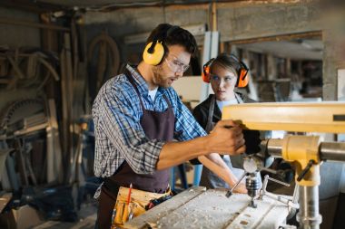 Usta woodworker kadın çırağı nasıl matkap basın, o onu dikkatle dinlerken kullanılacağını açıklayan göz ve kulak koruyucular