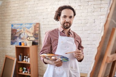 Art studio çalışma, poz ve kamera resim resim şövale üzerinde süre için gülümseyen yetenekli profesyonel sanatçı portresi