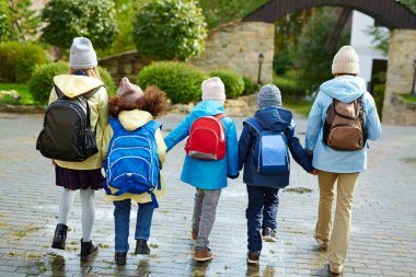 Schoolkids in casualwear with rucksacks behind backs holding by hands while going to school clipart