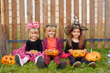 Youthful girls in halloween costumes sitting on grass by fence and eating treats clipart